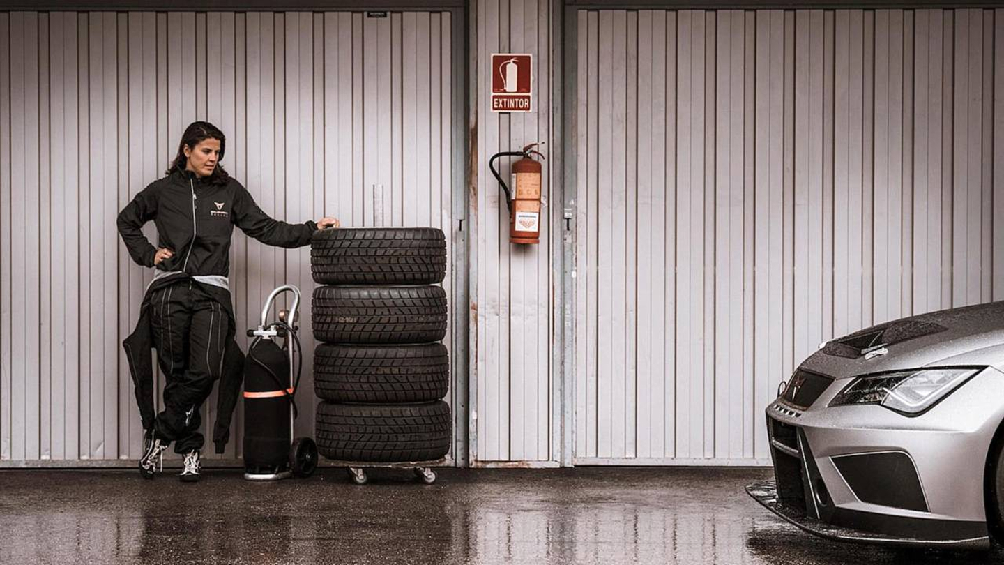 Frau in CUPRA Outfit lehnt an einem Stapel Autoreifen neben einem CUPRA Rennfahrzeug in einer Garage.